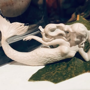 May include: A cream-colored mermaid figurine with black accents on the tail and hands. The mermaid is in a reclining pose, resting on a green and brown surface. The figurine is likely ceramic or porcelain.