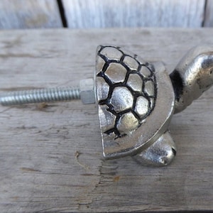 May include: A silver-colored turtle-shaped cabinet knob. The knob features a detailed turtle shell design and a protruding bolt for attachment. The hardware is made of metal and is designed for furniture.
