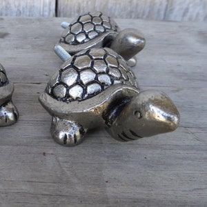May include: Four silver-colored turtle-shaped cabinet knobs. Each knob features a detailed shell pattern and a screw for attachment. The knobs are arranged on a weathered wooden surface, suggesting a rustic or nautical theme.