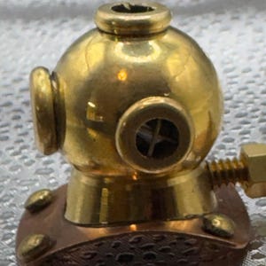 May include: Miniature brass diving helmet with copper base and threaded rod. The helmet has a round shape with portholes and a small top vent. A penny is next to the helmet for size comparison.