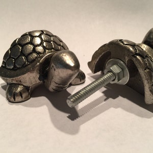 May include: Two silver-toned metal turtle cabinet knobs. The knobs are detailed with a textured shell design and have a bolt for attachment. One knob is fully assembled, while the other is partially disassembled, showing the bolt and washer.