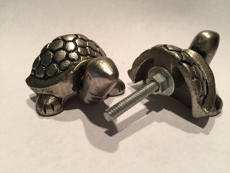 May include: Two silver-toned metal turtle cabinet knobs. The knobs are detailed with a textured shell design and have a bolt for attachment. One knob is fully assembled, while the other is partially disassembled, showing the bolt and washer.