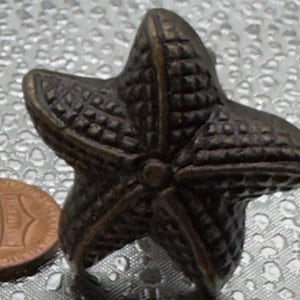 May include: Bronze-colored starfish-shaped cabinet knobs. The knobs have a textured surface and are next to a penny for size comparison. The knobs are likely for furniture or decorative use.