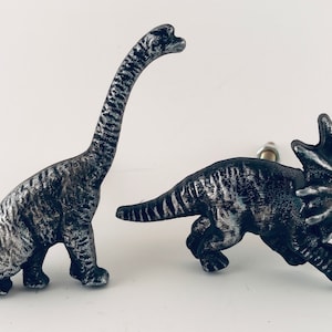 May include: Two dinosaur-shaped cabinet knobs. One is a long-necked Brachiosaurus, the other a Triceratops. Both are metallic silver and black, with detailed textures. These decorative knobs add a prehistoric touch to furniture.