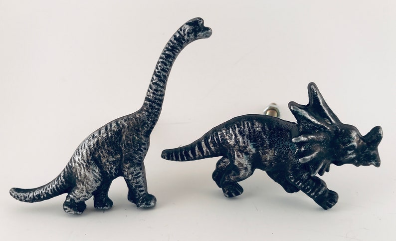 May include: Two dinosaur-shaped cabinet knobs. One is a long-necked Brachiosaurus, the other a Triceratops. Both are metallic silver and black, with detailed textures. These decorative knobs add a prehistoric touch to furniture.