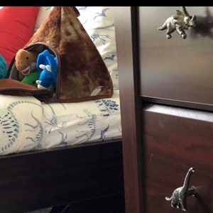 May include: A brown dinosaur-shaped storage bag filled with plush toy dinosaurs. The bag is on a bed with a dinosaur pattern. A dark brown chest of drawers with dinosaur-shaped drawer pulls is to the right.