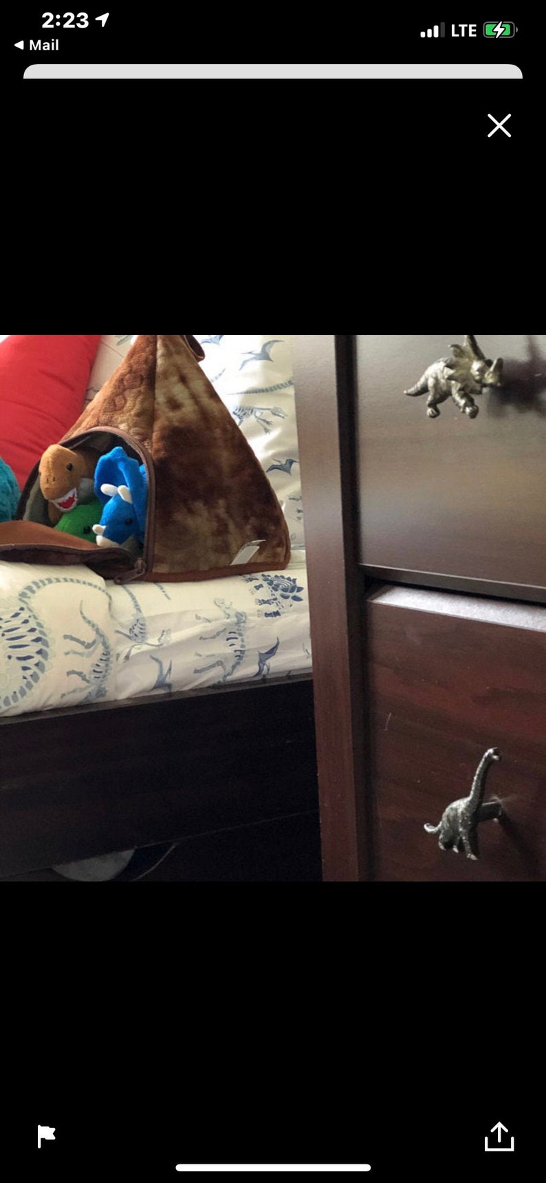 May include: A brown dinosaur-shaped storage bag filled with plush toy dinosaurs. The bag is on a bed with a dinosaur pattern. A dark brown chest of drawers with dinosaur-shaped drawer pulls is to the right.