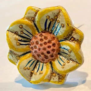 May include: A decorative flower-shaped cabinet knob. The knob features a yellow and off-white painted design with a pink center. The petals have yellow outlines and blue accents. Small gold-colored dots are scattered across the petals.