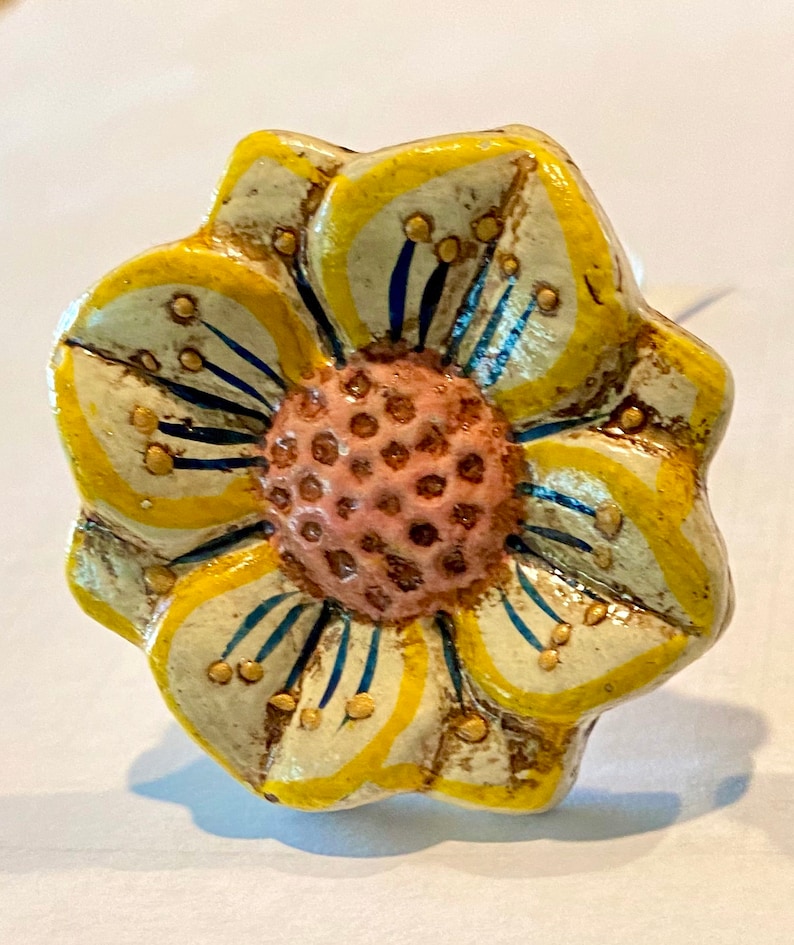 May include: A decorative flower-shaped cabinet knob. The knob features a yellow and off-white painted design with a pink center. The petals have yellow outlines and blue accents. Small gold-colored dots are scattered across the petals.