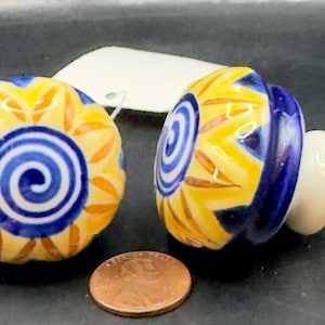 May include: Two decorative ceramic cabinet knobs. The knobs feature a yellow and blue floral design with a white base and a silver screw. A penny is next to the knobs for size comparison.