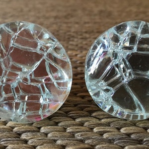 May include: Two clear, round glass cabinet knobs with a cracked ice design. The knobs are transparent, allowing light to pass through, and are set against a textured, neutral-toned background. These decorative knobs could be used for furniture or other DIY projects.