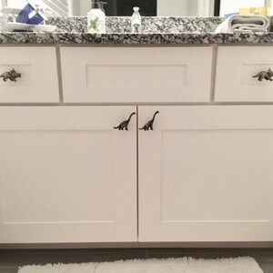 May include: White bathroom cabinet with dinosaur-shaped drawer pulls. The cabinet has two sets of drawers and doors. The drawer pulls are bronze-coloured and shaped like dinosaurs. The countertop is granite with a grey and white pattern.