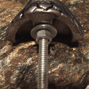 May include: Close-up of a silver-toned metal cabinet knob with a decorative design. The knob is attached to a textured, multi-colored stone surface with a bolt, washer, and nut.
