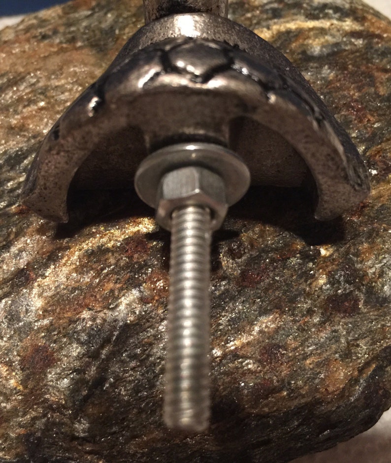 May include: Close-up of a silver-toned metal cabinet knob with a decorative design. The knob is attached to a textured, multi-colored stone surface with a bolt, washer, and nut.