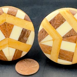 May include: Two round wooden cabinet knobs with a geometric design. The knobs feature a pattern of light brown, dark brown, and cream-colored wood pieces. A penny is next to the knobs for size comparison.