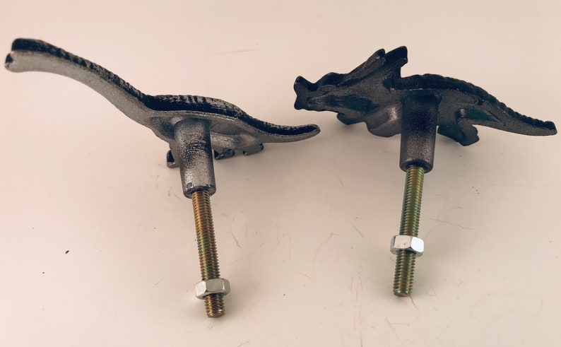 May include: Two cast iron dinosaur-shaped cabinet knobs. One is a long-necked Brontosaurus, the other a Triceratops. Both are painted in a distressed silver finish, with a threaded bolt and nut for mounting.