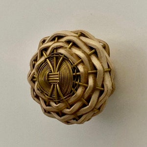 May include: A decorative cabinet knob with a woven, light beige exterior and a central brass-colored medallion. The knob has a rounded shape and is designed with a basket-weave pattern, offering a natural, textured appearance.