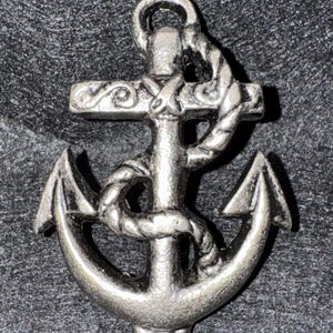 May include: A silver-colored anchor with a rope wrapped around the central shaft. The anchor's design includes a crossbar with decorative swirls and a curved top. The overall aesthetic is nautical.