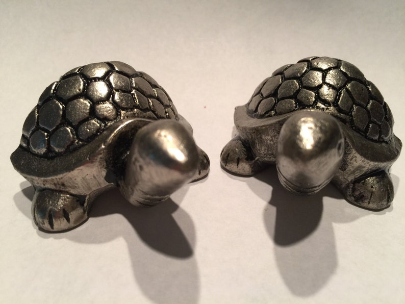 May include: Two silver-colored turtle figurines. Each turtle has a textured shell and detailed features. The turtles are positioned side-by-side, showcasing their front and side profiles. The figurines are likely decorative items.
