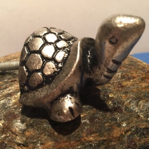 May include: A silver-colored metal turtle-shaped cabinet knob. The knob features a detailed shell design and a screw for attachment. The turtle's head is extended, and its face has etched details. This decorative hardware adds a whimsical touch to furniture.