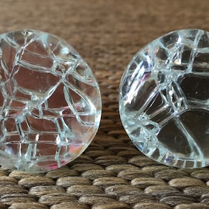 May include: Two clear, round glass cabinet knobs with a cracked ice design. The knobs are made of transparent glass and have a network of intersecting lines, creating a unique, textured appearance. The knobs are displayed on a woven surface.