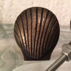 May include: Bronze-colored seashell cabinet knob with vertical ribbed detailing. The knob has a rounded, fan-like shape, and a small, square base. The hardware is designed for use on drawers or cabinet doors. The finish has a slightly aged appearance.