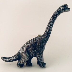 May include: A silver-coloured Brachiosaurus dinosaur figurine with a long neck and textured surface. The dinosaur is standing on a white surface. The figurine is a decorative item.
