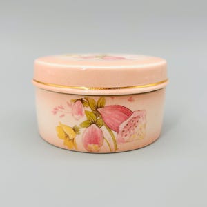 Peut inclure: Une boîte à bijoux ronde de couleur pêche avec un couvercle. La boîte est ornée d'un motif floral avec des fleurs roses et jaunes et des feuilles vertes. Une bande dorée entoure la boîte près du couvercle.