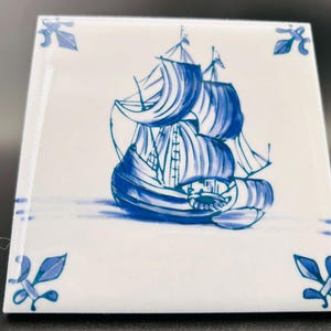 May include: Square ceramic tile with a hand-painted blue and white sailboat design. The nautical scene features a detailed ship with billowing sails. Decorative blue floral accents are in each corner. A classic, decorative piece.