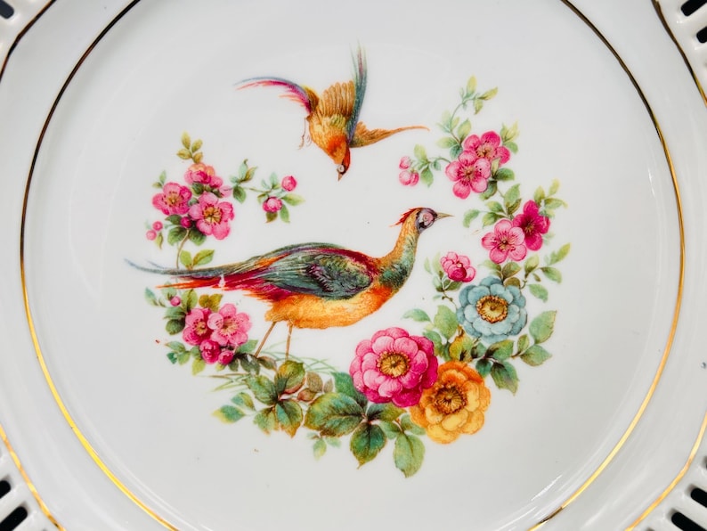 Peut inclure: Une assiette en porcelaine blanche avec un bord dor&eacute; pr&eacute;sente un motif peint &agrave; la main de deux oiseaux et de fleurs. Les oiseaux sont un faisan et un oiseau plus petit avec une poitrine jaune. Les fleurs sont roses, jaunes et bleues.