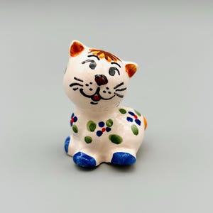 Peut inclure: Figurine de chat en céramique. La base est blanche, ornée de motifs floraux bleus, verts et rouges. Le chat a des oreilles oranges et un nez marron, avec des pieds bleus. Objet décoratif.