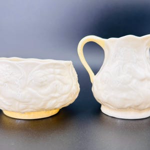 May include: A cream-colored ceramic creamer and sugar bowl set. The sugar bowl is round with a wide base and the creamer has a handle and spout. Both pieces have a floral embossed design and a yellow-toned base.