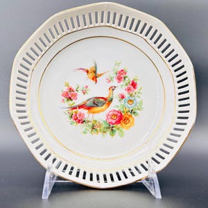 Peut inclure: Assiette en porcelaine blanche avec un bord dor&eacute; et un motif d&eacute;coup&eacute;. L'assiette pr&eacute;sente un motif floral avec deux oiseaux, un faisan et un oiseau plus petit, dans des tons de rouge, jaune et bleu.