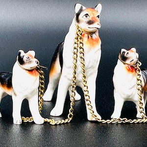 Peut inclure: Trois figurines de chiens en porcelaine avec des marques noires, blanches et brunes. Chaque chien porte un collier en chaîne dorée.