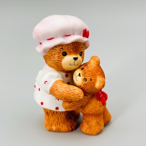Peut inclure: Figurine en céramique de deux ours en peluche s'embrassant. Le plus grand ours porte une robe blanche à pois rouges et un bonnet rose avec des accents rouges. Le plus petit ours a un nœud rouge. Les ours sont bruns avec des yeux et des nez noirs.