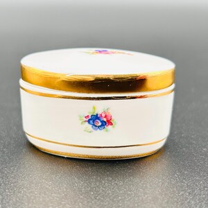 May include: A small, white ceramic trinket box with a gold rim and lid. The box is decorated with a floral design in shades of blue, pink, and yellow. The lid also features a floral design. The box is likely vintage or antique.