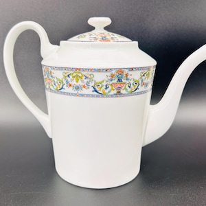 May include: White porcelain teapot with a curved handle and a long spout. The teapot features a decorative band with floral and geometric patterns in blue, orange, and green. The lid has a small knob and matching design.