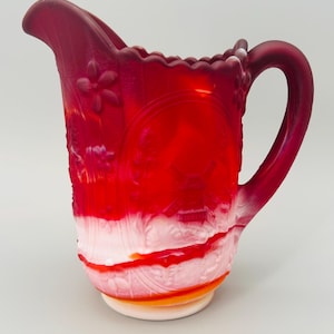 May include: A ruby red glass pitcher with a white base and handle. The pitcher features embossed floral and landscape designs. The rim has a scalloped edge. The pitcher is a decorative item.