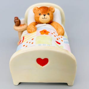 Peut inclure: Figurine en céramique d'un ours en peluche brun couché dans un lit crème avec un cœur rouge sur la tête de lit. L'ours est sous une courtepointe patchwork avec des motifs floraux et géométriques. Une petite table avec une bouteille et une tasse est à côté du lit.