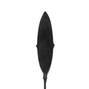Op de afbeelding: Een zwart, bladvormig serveerkeukengerei, mogelijk een spatel of serveerder, met een lange, dunne steel. Het keukengerei heeft een licht getextureerd oppervlak en een decoratief element in de buurt van de bovenkant van het blad. De achtergrond is effen wit.