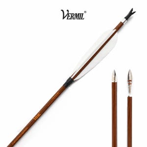 May include: A wooden archery arrow featuring white fletching, a black nock, and a silver-toned metal arrowhead. The shaft is brown with the text Vermil and Crafted for Champions printed in gold. A separate arrowhead is shown detached from a shaft.