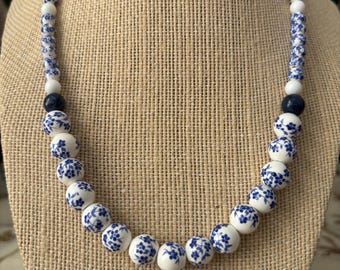 May include: A white ceramic necklace with blue floral designs. The necklace is made of round beads and has a simple design.