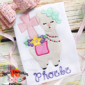 Puede incluir: Diseño bordado con una llama blanca con una cruz rosa, flores y un bolsillo rosa. La llama lleva una diadema verde y un collar de cuentas. El nombre "Phoebe" está bordado en morado debajo de la llama.