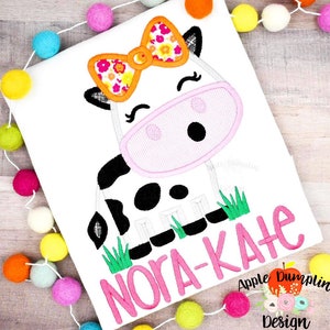 Puede incluir: Una vaca blanca con manchas negras que lleva un lazo rosa y naranja con estampado floral. La vaca est&aacute; de pie en la hierba verde. El nombre "Nora-Kate" est&aacute; escrito en letras rosas debajo de la vaca.