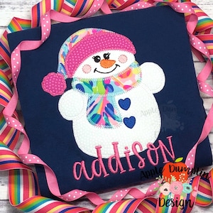 May include: A blue shirt with a white snowman applique. The snowman is wearing a pink hat, a colorful scarf, and has two blue hearts for buttons. The name "addison" is embroidered in pink below the snowman.