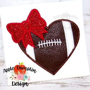 May include: A white background with a heart-shaped football design. The football is brown with white stripes and stitching. A red glitter bow is attached to the top left of the heart. The text "Apple Dumplin Design" is at the bottom.