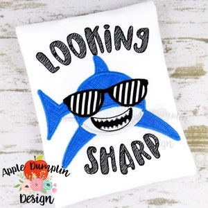 Puede incluir: Diseño bordado de un tiburón azul con gafas de sol y el texto "Looking Sharp" en negro.