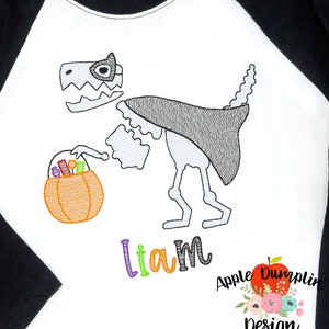 May include: A white t-shirt with a black and grey skeleton dinosaur wearing a cape and holding a pumpkin bucket with candy. The text "liam" is embroidered in colorful letters below the dinosaur. The shirt has a black and white baseball style sleeve.