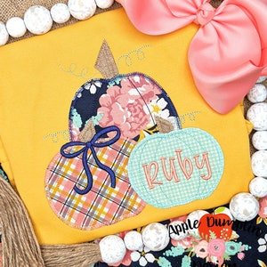 May include: A yellow shirt with a patch of three pumpkins. The pumpkins are made of fabric with different patterns. The largest pumpkin is a floral pattern, the middle pumpkin is a plaid pattern, and the smallest pumpkin is a solid green color with the name "Ruby" embroidered on it.