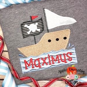 Puede incluir: Una camiseta gris con un diseño bordado de un barco pirata con una bandera de calavera y huesos cruzados. El barco navega en aguas azules con olas blancas. El nombre "MAXIMUS" está bordado en rojo en una pancarta a rayas azules y blancas debajo del barco.